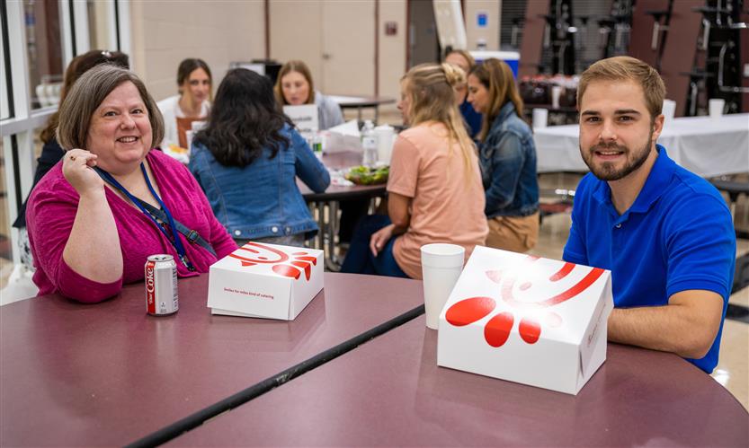A man and woman sit at a cafeteria tables with lunch boxes in front of them